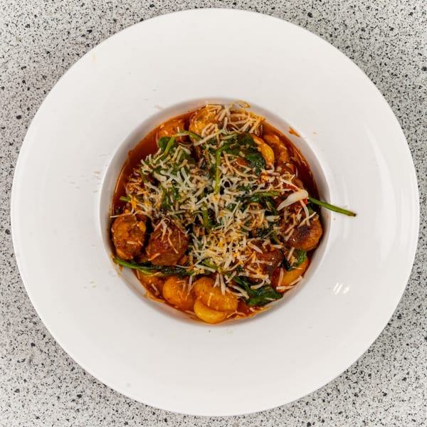 A white bowl filled with gnocchi, meatballs, spinach, and grated cheese in a red sauce.