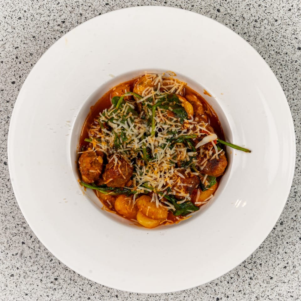 A white bowl filled with gnocchi, meatballs, spinach, and grated cheese in a red sauce.