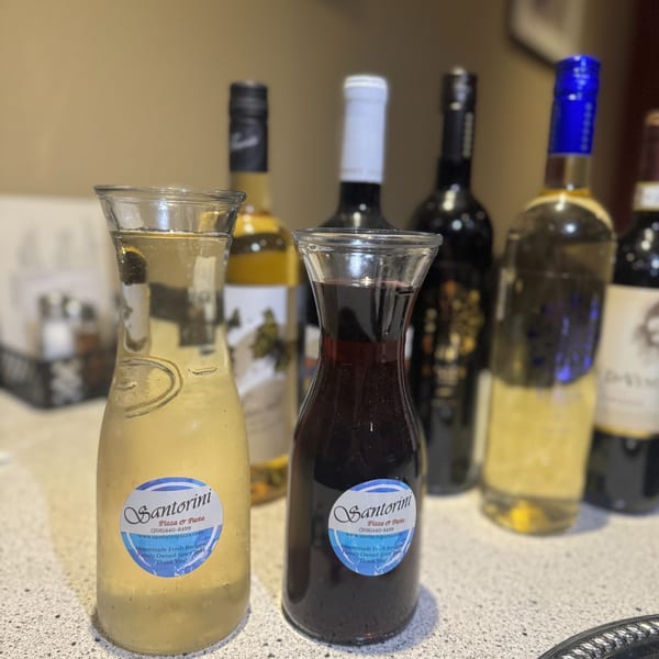 Two carafes of Santorini Pizza & Pasta salad dressing, with wine bottles blurred in the background.