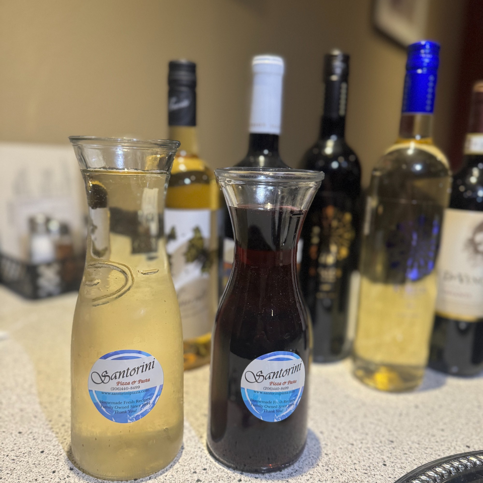 Two carafes of Santorini Pizza & Pasta salad dressing, with wine bottles blurred in the background.