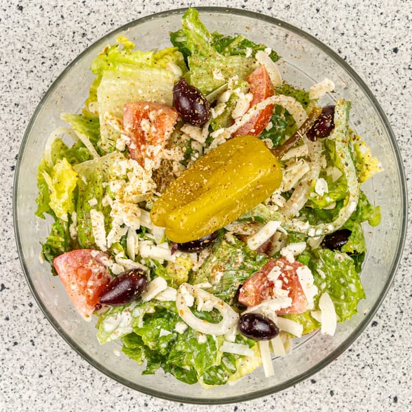 A vibrant Greek salad in a clear bowl, featuring lettuce, tomatoes, olives, feta, and a pepperoncini.